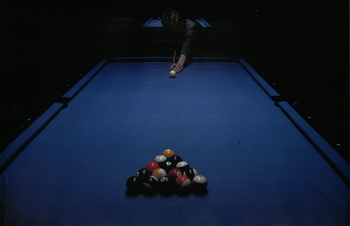 Photograph of a figure in front of a pool table, preparing to play, with pool balls in foreground